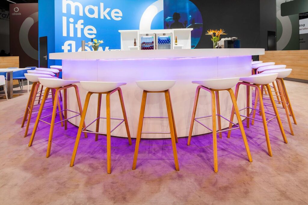 White bar stools with oak legs around a curved lit bar on a stand at ISH 2019 in Frankfurt