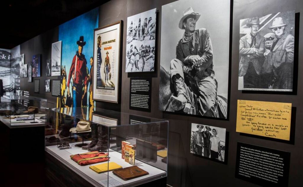 John Wayne exhibit with framed photos, film poster and glass display cases of hats and boots
