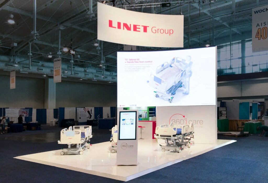 LINET Group booth with curved hanging sign, large backlit graphic wall and hospital beds on white floor