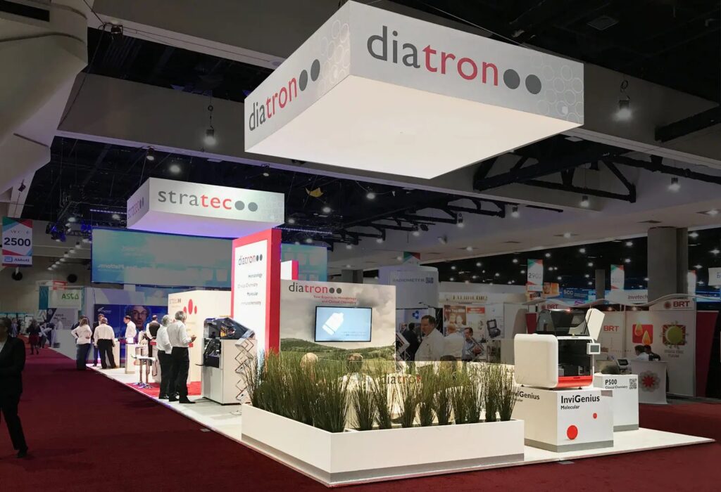 Diatron and Stratec island booths with hanging signs, planters, lab analysers and visitors in the aisle