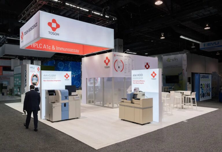 Tosoh Bioscience island booth with hanging red and white sign, lit wall panels and two immunoassay analysers