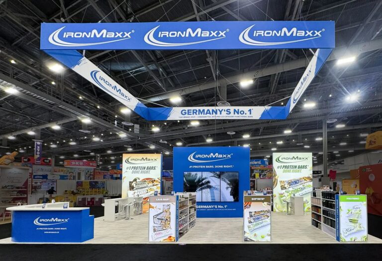 IronMaxx island booth with blue hanging banner, protein bar displays, shelving and video wall