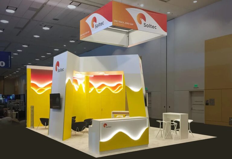 Soltec booth with yellow wave-shaped backlit walls, white counter, lounge chairs and orange hanging sign