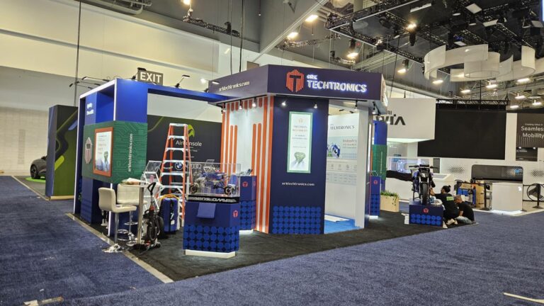 Blue and orange SRK Techtronics stand during final set-up, with display plinths and a motorbike exhibit