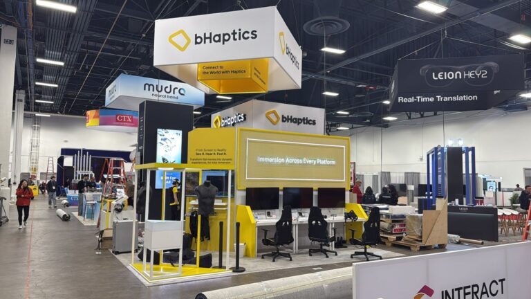 Yellow and white bHaptics stand at CES 2026 with hanging square banner, gaming chairs and screen wall