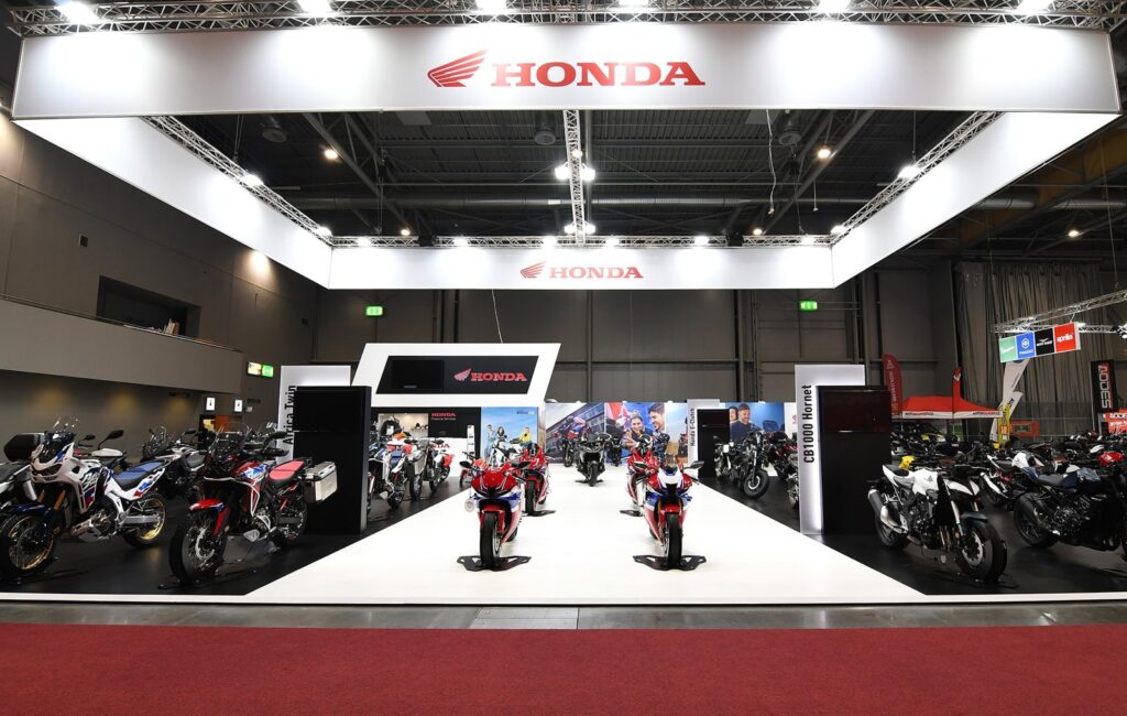 Honda motorcycle stand under a white suspended banner with bikes along a white central aisle at Motosalon Brno